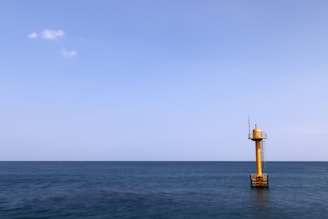 A solitary yellow buoy tower is positioned in the vast expanse of the ocean, under a clear blue sky. The sea is calm, and a few small clouds are scattered in the sky.