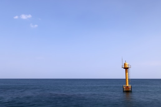 A solitary yellow buoy tower is positioned in the vast expanse of the ocean, under a clear blue sky. The sea is calm, and a few small clouds are scattered in the sky.