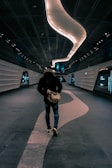 A person is walking down a futuristic, dimly-lit corridor with sleek, metallic walls and a wavy light fixture on the ceiling. The individual is wearing a jacket and jeans, carrying a bag, and the hallway gives off a modern, sci-fi ambiance.