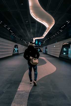 A person is walking down a futuristic, dimly-lit corridor with sleek, metallic walls and a wavy light fixture on the ceiling. The individual is wearing a jacket and jeans, carrying a bag, and the hallway gives off a modern, sci-fi ambiance.