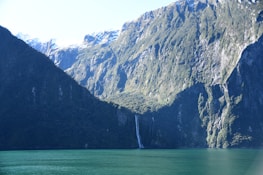 A stunning natural landscape featuring a large, rugged mountain range with steep cliffs and a waterfall cascading down the side. The mountains are partially covered in snow, and lush green vegetation is visible on the slopes. At the base of the mountains lies a deep turquoise lake under a clear blue sky.