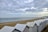 A row of white beach huts is lined up along a sandy beach. The beach stretches out towards the calm ocean, which extends to the horizon. A few people can be seen walking along the shore. The sky is mostly covered with clouds, with patches of blue peeking through.