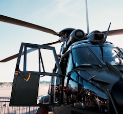 A close-up view of a black military helicopter with its door open. The helicopter features large rotor blades and several mechanical components visible around the top. The background shows an airfield with some infrastructure in the distance under a clear blue sky.