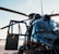 A close-up view of a black military helicopter with its door open. The helicopter features large rotor blades and several mechanical components visible around the top. The background shows an airfield with some infrastructure in the distance under a clear blue sky.