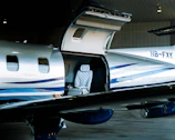 A sleek, modern private jet with an open hatch is parked inside a hangar. Visible through the hatch is a luxurious interior featuring a quilted leather seat. The exterior is silver with blue stripes, and the tail number HB-FXK can be seen on the fuselage.