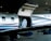 A sleek, modern private jet with an open hatch is parked inside a hangar. Visible through the hatch is a luxurious interior featuring a quilted leather seat. The exterior is silver with blue stripes, and the tail number HB-FXK can be seen on the fuselage.