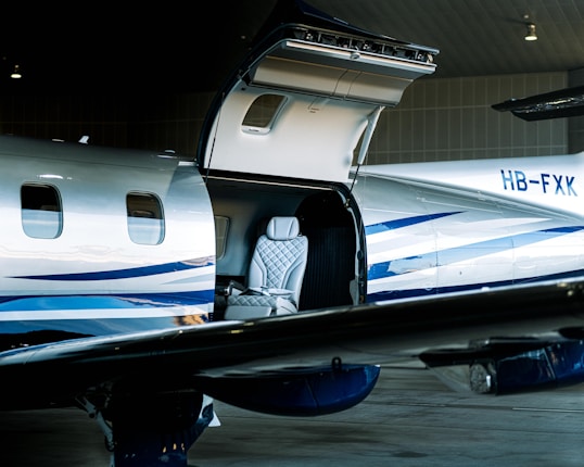 A sleek, modern private jet with an open hatch is parked inside a hangar. Visible through the hatch is a luxurious interior featuring a quilted leather seat. The exterior is silver with blue stripes, and the tail number HB-FXK can be seen on the fuselage.