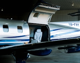 A sleek, modern private jet with an open hatch is parked inside a hangar. Visible through the hatch is a luxurious interior featuring a quilted leather seat. The exterior is silver with blue stripes, and the tail number HB-FXK can be seen on the fuselage.