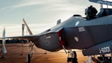 A modern fighter jet parked on an airfield in the evening light. Various warning labels and technical markings are visible on the aircraft's body. The surrounding area has blue banners and orange tarmac, with a metal fence in the background.