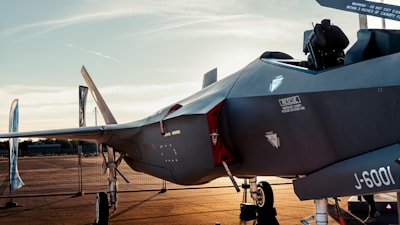 A modern fighter jet parked on an airfield in the evening light. Various warning labels and technical markings are visible on the aircraft's body. The surrounding area has blue banners and orange tarmac, with a metal fence in the background.