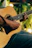 A focused man playing guitar outdoors with sunlight filtering through trees.