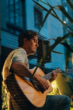 A man is seated indoors, playing an acoustic guitar. The setting appears to be relaxed with some greenery in the foreground. The lighting is soft, creating a calm ambiance, and the background includes vertical blinds suggesting a window.