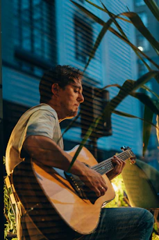 A man is seated indoors, playing an acoustic guitar. The setting appears to be relaxed with some greenery in the foreground. The lighting is soft, creating a calm ambiance, and the background includes vertical blinds suggesting a window.