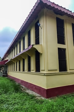 A long, two-story yellow building with a maroon base and roof. The building has multiple black-barred windows along the side and is surrounded by green grass. The roof has a distinct wavy design, and there is a security light installed on the corner of the building.