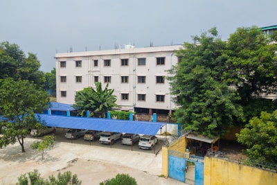 A four-story beige building surrounded by lush green trees. In front of the building, there is a parking area with several parked cars under a blue metal canopy. A banana plant is noticeable near the entrance. The scene is quiet and serene with a clear sky overhead.