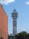 A tall tower with a modern design features the large vertical red letters spelling 'CASINO'. The structure has a sleek, metallic appearance with various horizontal and vertical lines. The sky is mostly clear with a few clouds. In the foreground, part of a brick building is visible, along with some trees and buildings in the distance.