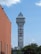 A tall tower with a modern design features the large vertical red letters spelling 'CASINO'. The structure has a sleek, metallic appearance with various horizontal and vertical lines. The sky is mostly clear with a few clouds. In the foreground, part of a brick building is visible, along with some trees and buildings in the distance.