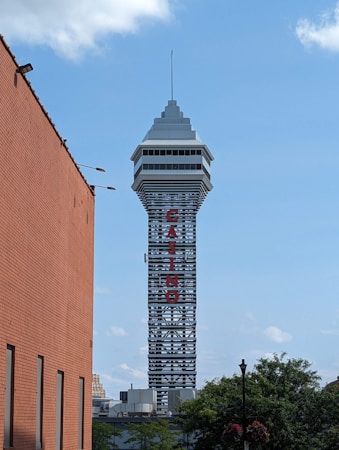 A tall tower with a modern design features the large vertical red letters spelling 'CASINO'. The structure has a sleek, metallic appearance with various horizontal and vertical lines. The sky is mostly clear with a few clouds. In the foreground, part of a brick building is visible, along with some trees and buildings in the distance.