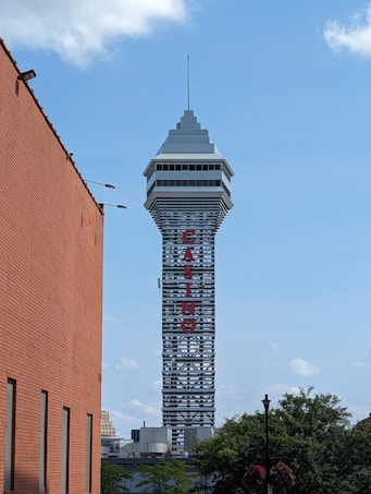 A tall tower with a modern design features the large vertical red letters spelling 'CASINO'. The structure has a sleek, metallic appearance with various horizontal and vertical lines. The sky is mostly clear with a few clouds. In the foreground, part of a brick building is visible, along with some trees and buildings in the distance.