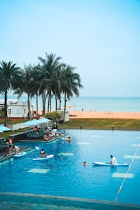 A lively scene of a swimming pool with people enjoying various activities. Several individuals are on paddleboards, while others are swimming and lounging by the poolside. Tall palm trees are visible in the background, and beyond them, there's a view of a sandy beach meeting the calm, blue ocean.
