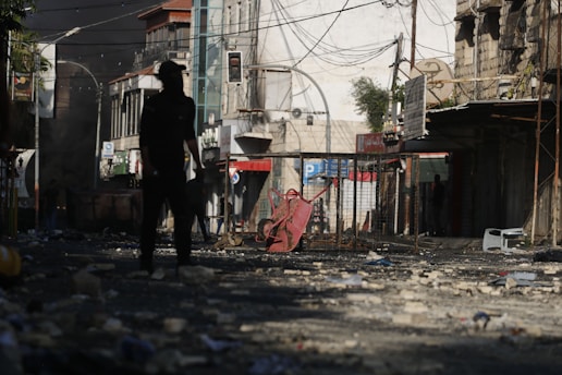 A street is littered with debris and rubble. In the foreground, a silhouette of a person is visible, standing amidst the destruction. The background features damaged buildings, with wires hanging and signs of disrepair. Smoke appears to be rising from the area, indicating a recent disturbance or unrest.