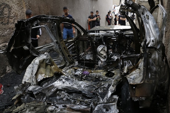 A burnt-out car is situated in a narrow alleyway, with several people standing nearby, some taking photos. The car's metal frame is warped and charred black, indicating severe damage. The surrounding walls are seen in the background.