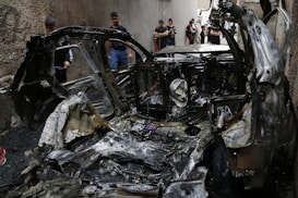 A burnt-out car is situated in a narrow alleyway, with several people standing nearby, some taking photos. The car's metal frame is warped and charred black, indicating severe damage. The surrounding walls are seen in the background.