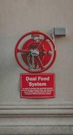 A large red control valve mounted on a neutral-colored wall, with a metal chain attached to it. Below the valve, a red sign reads 'Dual Feed System' and provides instructions for isolating the sprinkler system.