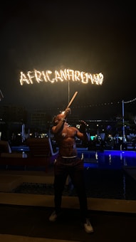 A fire performer is standing outdoors at night, skillfully manipulating a flaming baton. The word 'AFRICANFIREMAN' is emblazoned in flames above, illuminating the dark surroundings. The setting appears to be near a poolside with loungers and subtle lighting, creating a vibrant atmosphere.