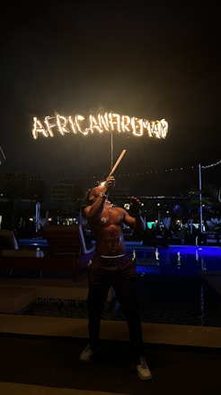 A fire performer is standing outdoors at night, skillfully manipulating a flaming baton. The word 'AFRICANFIREMAN' is emblazoned in flames above, illuminating the dark surroundings. The setting appears to be near a poolside with loungers and subtle lighting, creating a vibrant atmosphere.
