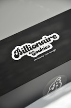 A sleek black box features the logo 'Billionaire Cookies' in a bold, stylized font, accompanied by the tagline 'seriously rich dough' in smaller letters. The box has a minimalistic design with a focus on the company branding.
