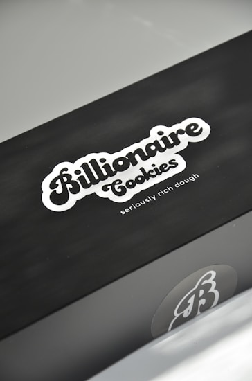A sleek black box features the logo 'Billionaire Cookies' in a bold, stylized font, accompanied by the tagline 'seriously rich dough' in smaller letters. The box has a minimalistic design with a focus on the company branding.