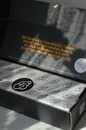 The image features a partly open black box with branded wrapping paper inside. The interior of the lid displays an eye-catching text about cookies: 'Bil•ion•aire Coo•kie(s) - Noun: Hand-crafted cookie dough so rich in flavor it's like your tastebuds struck gold.' Natural light creates soft shadows, adding depth to the scene.