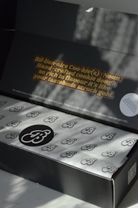 The image features a partly open black box with branded wrapping paper inside. The interior of the lid displays an eye-catching text about cookies: 'Bil•ion•aire Coo•kie(s) - Noun: Hand-crafted cookie dough so rich in flavor it's like your tastebuds struck gold.' Natural light creates soft shadows, adding depth to the scene.