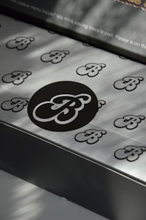 A pattern of stylized 'B' letters is printed on a smooth metallic or paper surface. A large black circle in the center highlights one of the 'B' letters, with smaller ones scattered around it. There's a message at the top regarding saving trees, indicating an environmentally conscious theme.