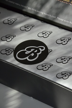 A pattern of stylized 'B' letters is printed on a smooth metallic or paper surface. A large black circle in the center highlights one of the 'B' letters, with smaller ones scattered around it. There's a message at the top regarding saving trees, indicating an environmentally conscious theme.