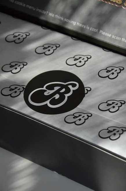 A pattern of stylized 'B' letters is printed on a smooth metallic or paper surface. A large black circle in the center highlights one of the 'B' letters, with smaller ones scattered around it. There's a message at the top regarding saving trees, indicating an environmentally conscious theme.
