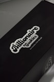 A black box with a white logo of the brand 'Billionaire Cookies', featuring a bold, cursive font. The tagline 'seriously rich dough' is printed underneath. The overall appearance is sleek and minimalistic.