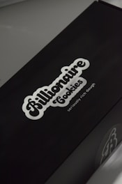 A black box with a white logo of the brand 'Billionaire Cookies', featuring a bold, cursive font. The tagline 'seriously rich dough' is printed underneath. The overall appearance is sleek and minimalistic.