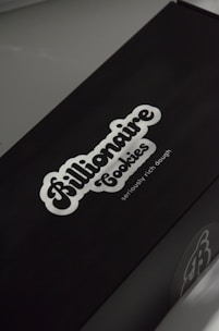 A black box with a white logo of the brand 'Billionaire Cookies', featuring a bold, cursive font. The tagline 'seriously rich dough' is printed underneath. The overall appearance is sleek and minimalistic.