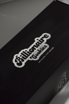 A black box with a white logo of the brand 'Billionaire Cookies', featuring a bold, cursive font. The tagline 'seriously rich dough' is printed underneath. The overall appearance is sleek and minimalistic.