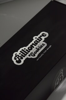 A black box with a white logo of the brand 'Billionaire Cookies', featuring a bold, cursive font. The tagline 'seriously rich dough' is printed underneath. The overall appearance is sleek and minimalistic.