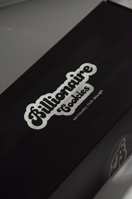 A black box with a white logo of the brand 'Billionaire Cookies', featuring a bold, cursive font. The tagline 'seriously rich dough' is printed underneath. The overall appearance is sleek and minimalistic.