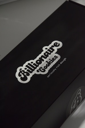 A black box with a white logo of the brand 'Billionaire Cookies', featuring a bold, cursive font. The tagline 'seriously rich dough' is printed underneath. The overall appearance is sleek and minimalistic.