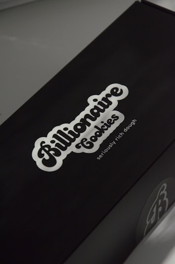 A black box with a white logo of the brand 'Billionaire Cookies', featuring a bold, cursive font. The tagline 'seriously rich dough' is printed underneath. The overall appearance is sleek and minimalistic.