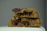 A close-up view of a chocolate chip cookie, broken in half, stacked on a surface. The cookie appears to be soft with large, melted chocolate chunks visible.