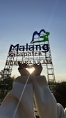 A large sign with the words 'Malang Kabupaten' stands against a clear blue sky. It features a stylized M in blue and green. Below it, someone creates a heart shape with their hands, framing the bright sun, which casts a warm glow around their fingers. The area is surrounded by greenery and some small plants in the foreground.