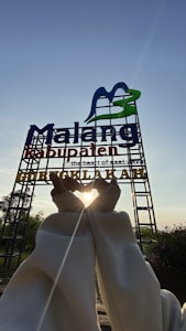 A large sign with the words 'Malang Kabupaten' stands against a clear blue sky. It features a stylized M in blue and green. Below it, someone creates a heart shape with their hands, framing the bright sun, which casts a warm glow around their fingers. The area is surrounded by greenery and some small plants in the foreground.