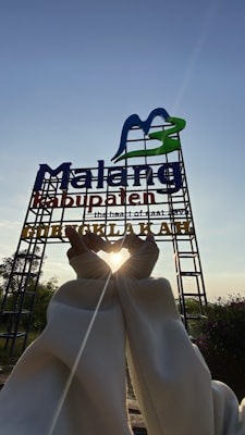 A large sign with the words 'Malang Kabupaten' stands against a clear blue sky. It features a stylized M in blue and green. Below it, someone creates a heart shape with their hands, framing the bright sun, which casts a warm glow around their fingers. The area is surrounded by greenery and some small plants in the foreground.