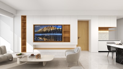 A modern living room features minimalistic design elements with a light color palette. A large flat-screen TV is mounted on a wall with built-in white shelving units, accented with wooden panels. The room is furnished with contemporary chairs and tables. Sunlight streams through a window, casting soft shadows on the polished floor.